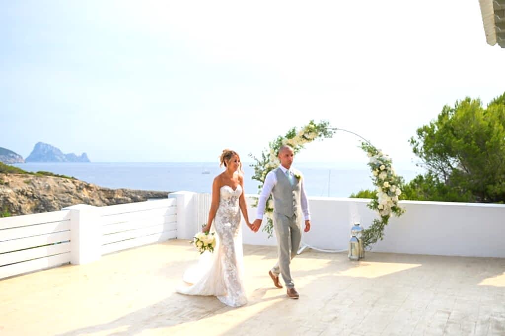 A bride and groom holding hands and walking on a terrace overlooking the sea, with a floral arch in the background. The groom is in a light grey suit, and the bride is in a strapless white lace wedding dress.