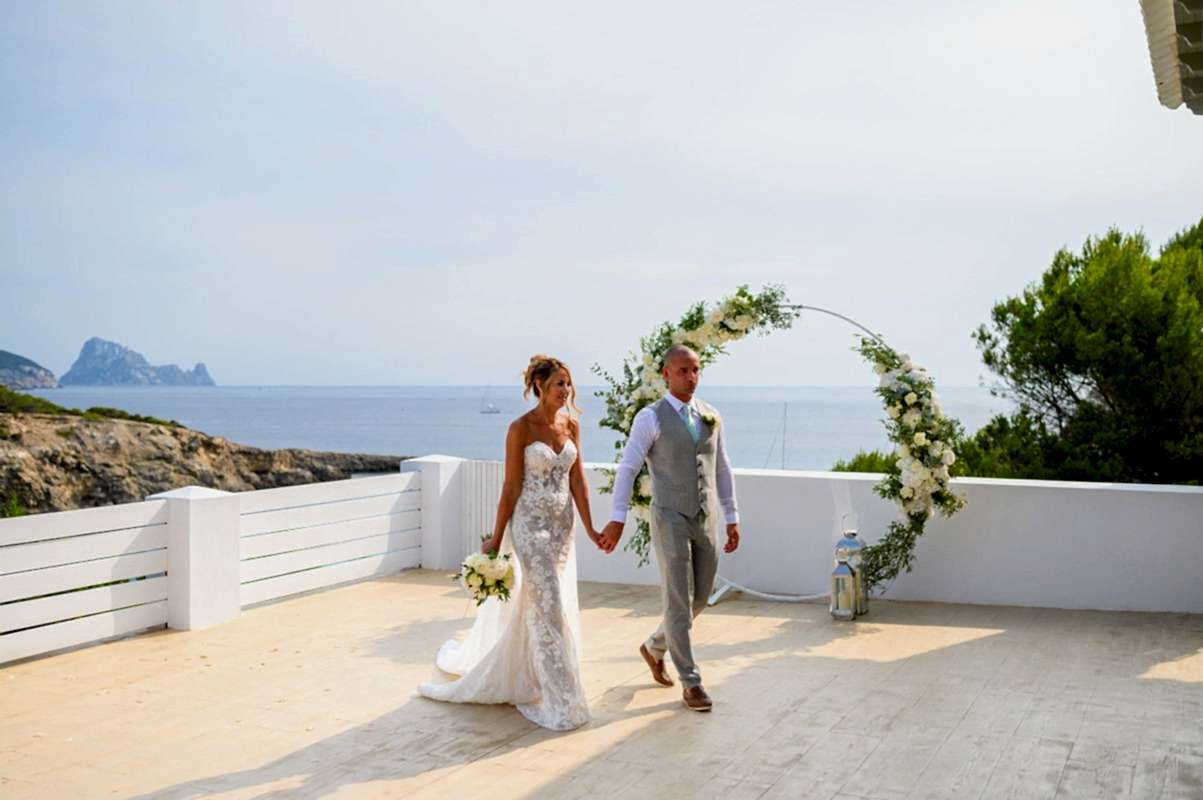 A bride and groom holding hands and walking on a terrace overlooking the sea, with a floral arch in the background. The groom is in a light grey suit, and the bride is in a strapless white lace wedding dress.
